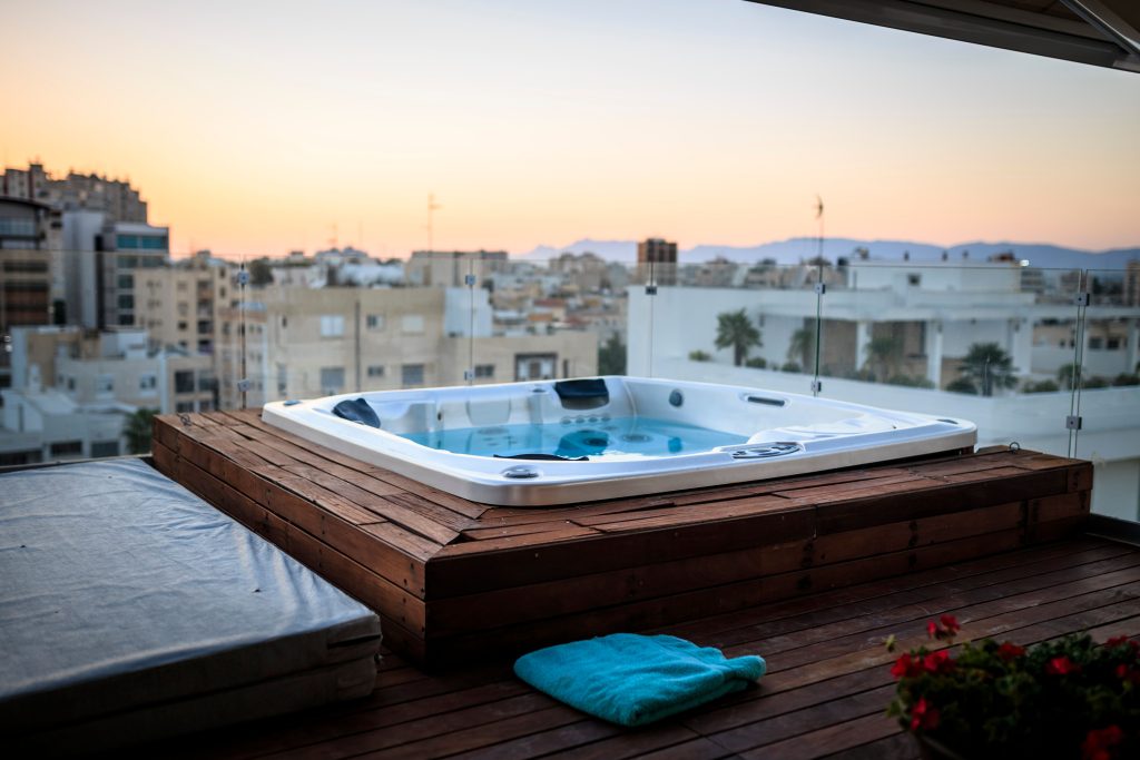 A square white hot tub with a folded insulated cover on a wooden rooftop terrace facing the city skyline